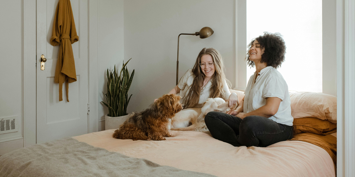 twee vrouwen die samen met een hond op bed zitten en lachen.